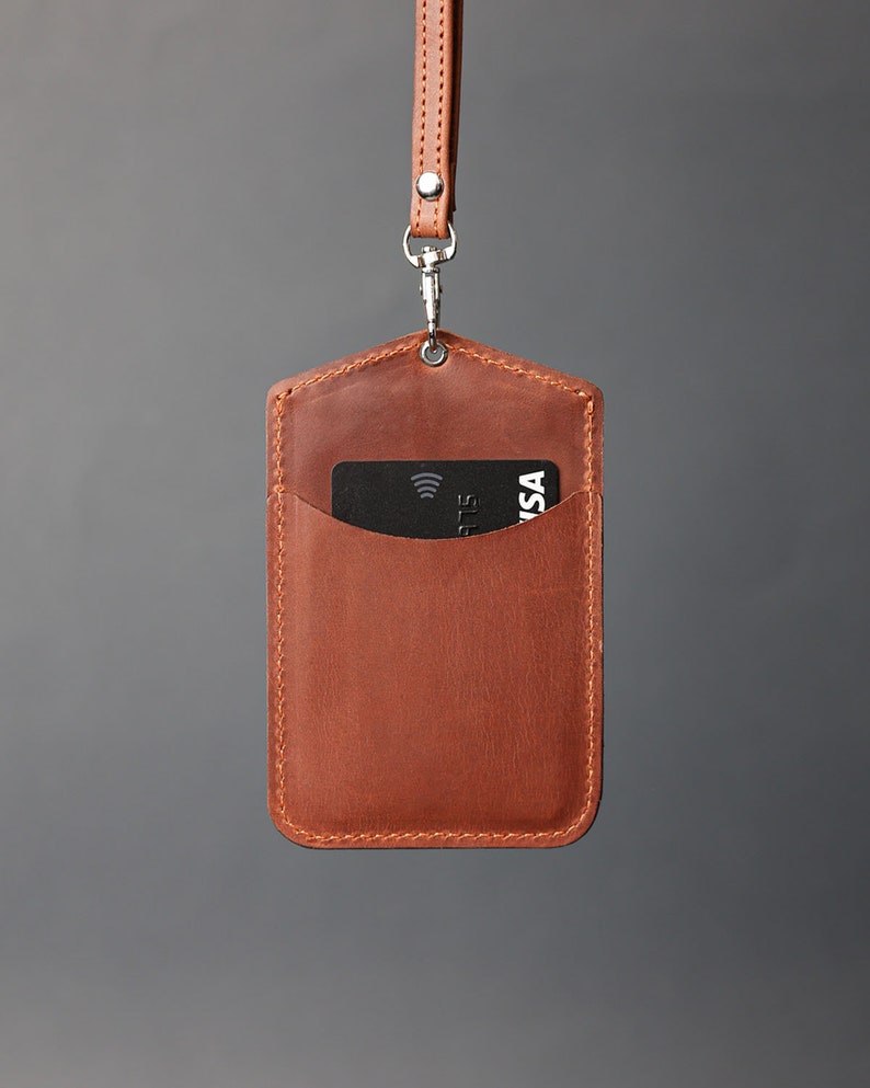 May include: A brown leather ID badge holder with a black card inside. The holder has a stitched edge and a silver chain attached to the top.