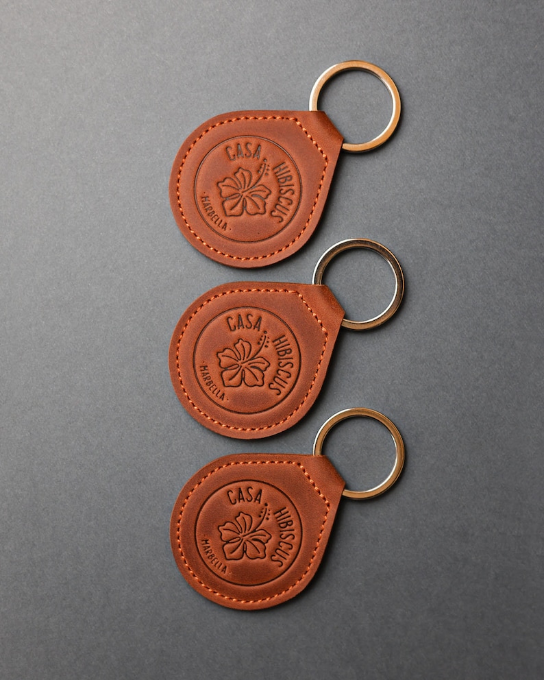 May include: Three brown leather keychains with a silver key ring. Each keychain features a circular design with the text "Casa Hibiscus Marbella" and a stylized hibiscus flower.