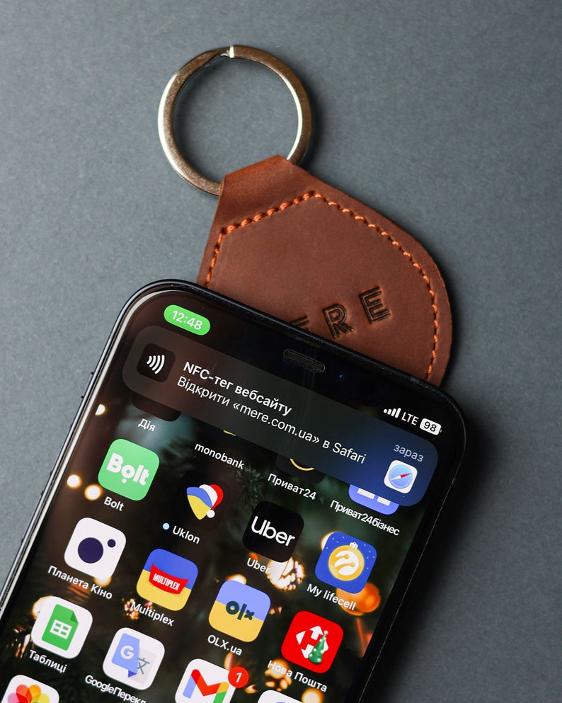 May include: A brown leather keychain with the word "KRE" embossed on it. The keychain is attached to a smartphone with a screen displaying various app icons, including Uber, Bolt, and Privat24. The phone screen also shows a notification for an NFC tag website.