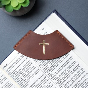 May include: A brown leather bookmark with a gold cross design. The bookmark is placed on an open book with text in a foreign language.