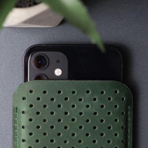 May include: A dark green leather phone sleeve with a perforated design. The sleeve is partially covering a black smartphone, with the camera lenses visible. The background is a dark gray surface, with a plant in a white pot in the upper left corner.