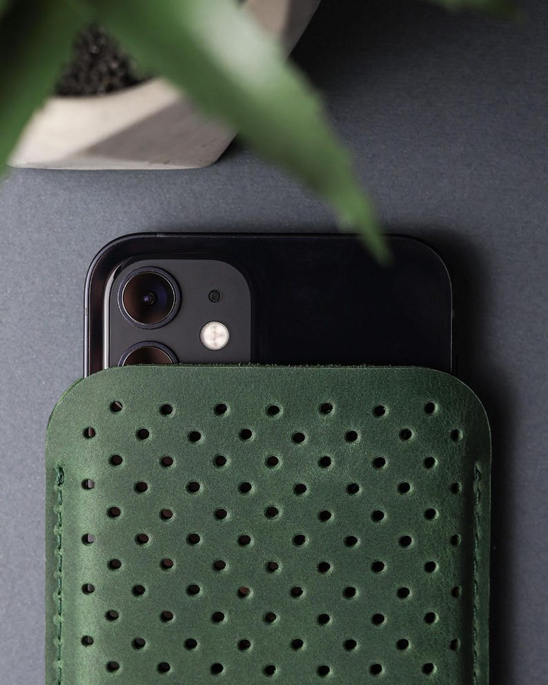 May include: A dark green leather phone sleeve with a perforated design. The sleeve is partially covering a black smartphone, with the camera lenses visible. The background is a dark gray surface, with a plant in a white pot in the upper left corner.