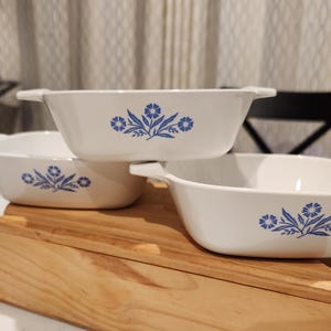 Könnte beinhalten: Drei weiße Keramik-Auflaufformen mit blauen Blumenmustern. Die Auflaufformen sind auf einem Serviertablett aus Holz gestapelt. Jede Form hat einen Griff auf beiden Seiten. Das Blumenmuster ist eine stilisierte blaue Blume mit Blättern.