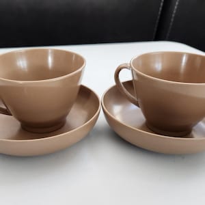 May include: Two matching tan-colored teacups and saucers. The cups have a rounded shape with a handle, and the saucers are a similar color and shape. The set is placed on a white surface.