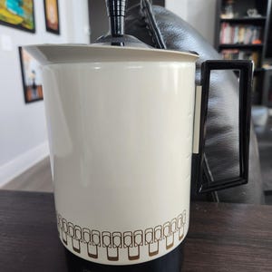 Puede incluir: Cafetera Empire Supreme de color crema con asa, tapa y base negras. La cafetera tiene un patrón decorativo marrón alrededor de la parte inferior y el logotipo de Empire Supreme. La cafetera está sobre una superficie marrón oscuro.