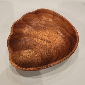 May include: A wooden bowl with a unique, organic shape. The bowl is made of a warm, medium-toned wood, showcasing the natural grain patterns. The edges have a scalloped design, adding a decorative touch. This bowl is suitable for serving snacks or as a decorative piece.