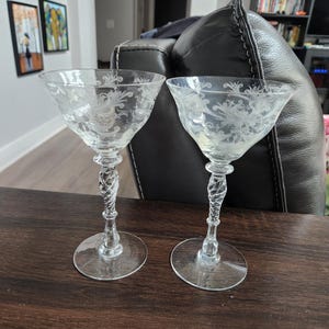 Puede incluir: Dos copas de champán vintage transparentes con diseños florales grabados. Los tallos son retorcidos y las bases son redondas. Las copas se exhiben sobre una superficie de madera marrón oscuro.