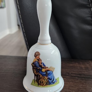 Puede incluir: Campana de porcelana blanca con un mango largo y un borde dorado. La campana presenta una escena pintada de una persona leyendo a un niño. La persona lleva un vestido azul y el niño un vestido amarillo. El objeto es decorativo.