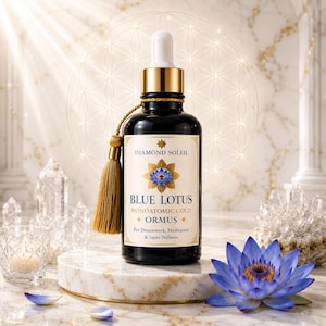 May include: A dark blue glass bottle with a gold dropper and tassel, labeled "Blue Lotus Monoatomic Gold + Ormus." The label features a blue lotus flower illustration. The bottle sits on a marble stand, with a blue lotus flower and crystals nearby.