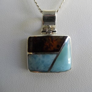 May include: A silver pendant with a square-shaped stone inlay. The stone is a mix of brown, blue, and white.