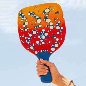 Puede incluir: Una paleta de playa de madera con un mango azul y una cara colorida. La cara de la paleta tiene un degradado de naranja a rojo con diseños blancos y azules en forma de burbujas. La palabra "CHAMPION" está impresa en la paleta.