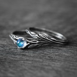 Könnte beinhalten: Zwei silberne Ringe mit einem von der Natur inspirierten Design. Ein Ring hat einen runden, blauen Edelstein. Die Bänder haben ein detailliertes, blattartiges Muster. Die Ringe werden auf einer dunklen, strukturierten Oberfläche präsentiert.