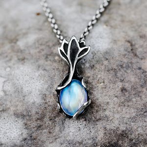 May include: A silver-toned pendant necklace featuring a blue and white oval gemstone. The pendant has a stylized floral design and hangs from a delicate chain. The gemstone has a pearlescent sheen.
