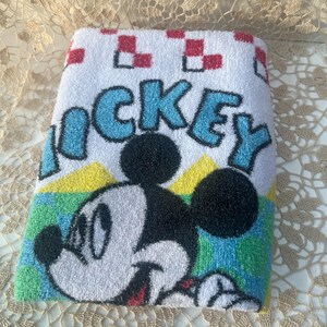 May include: Folded white towel with a Mickey Mouse design. The word "MICKEY" is in blue letters. Mickey Mouse is smiling, with black ears and head, and a white face. The towel has a red and white checkered pattern.