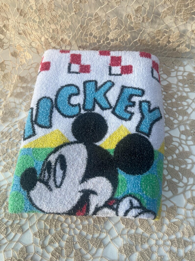 May include: Folded white towel with a Mickey Mouse design. The word "MICKEY" is in blue letters. Mickey Mouse is smiling, with black ears and head, and a white face. The towel has a red and white checkered pattern.