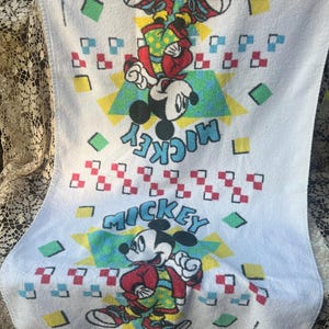 May include: White Mickey Mouse towel with two images of Mickey Mouse in red, yellow, and green outfits. The word "Mickey" is written in turquoise. The towel has a geometric pattern of squares and triangles.