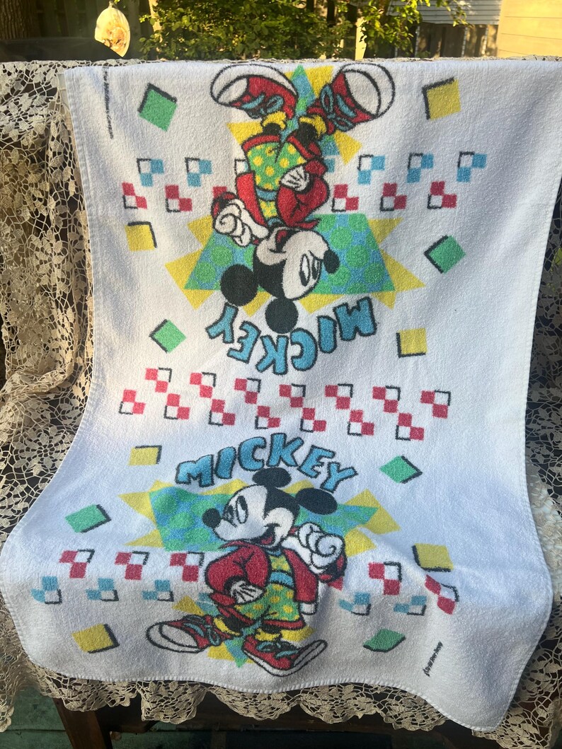 May include: White Mickey Mouse towel with two images of Mickey Mouse in red, yellow, and green outfits. The word "Mickey" is written in turquoise. The towel has a geometric pattern of squares and triangles.