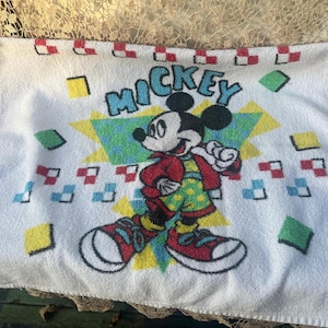 May include: White rectangular towel with a Mickey Mouse design. The graphic shows Mickey in a red jacket and green polka-dot shorts, with the word "MICKEY" in turquoise. The design is surrounded by colorful geometric shapes.