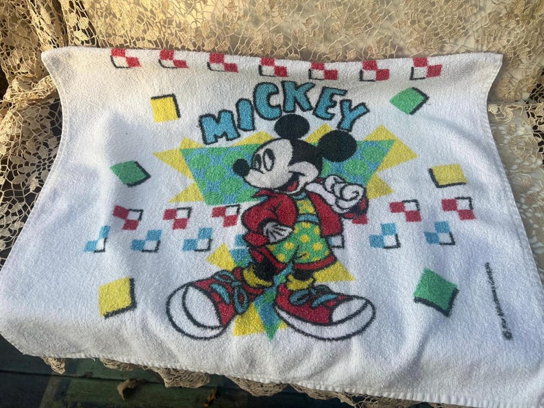 May include: White rectangular towel with a Mickey Mouse design. The graphic shows Mickey in a red jacket and green polka-dot shorts, with the word "MICKEY" in turquoise. The design is surrounded by colorful geometric shapes.