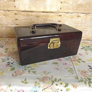 May include: A dark red, rectangular, plastic jewelry box with a gold latch and handle. The box has a glossy finish and is sitting on a floral tablecloth.
