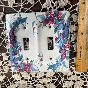 May include: A white ceramic light switch cover with a floral design featuring butterflies in shades of blue, purple, and pink. The design is surrounded by a border of green leaves and red flowers.