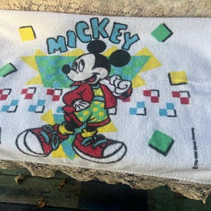 May include: White towel with a Mickey Mouse design. Mickey is wearing a red jacket, green polka-dot shirt, and red trainers. The word "Mickey" is in blue letters. Geometric shapes in various colours surround the character.