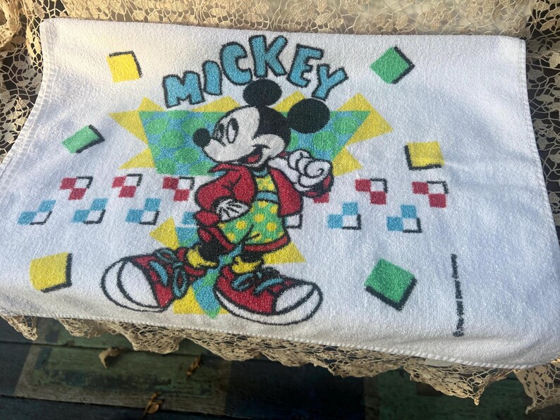 May include: White towel with a Mickey Mouse design. Mickey is wearing a red jacket, green polka-dot shirt, and red trainers. The word "Mickey" is in blue letters. Geometric shapes in various colours surround the character.