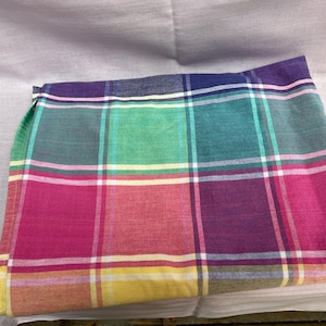 May include: A folded fabric textile featuring a multi-colored plaid pattern. The design consists of intersecting horizontal and vertical lines in shades of magenta, teal, purple, yellow, and white, creating a grid of various colored squares.