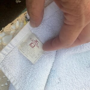May include: White Cannon brand towel with a label visible. The label has the Cannon logo and text. The towel is folded and held by a hand. The towel is on top of a beige crocheted surface.