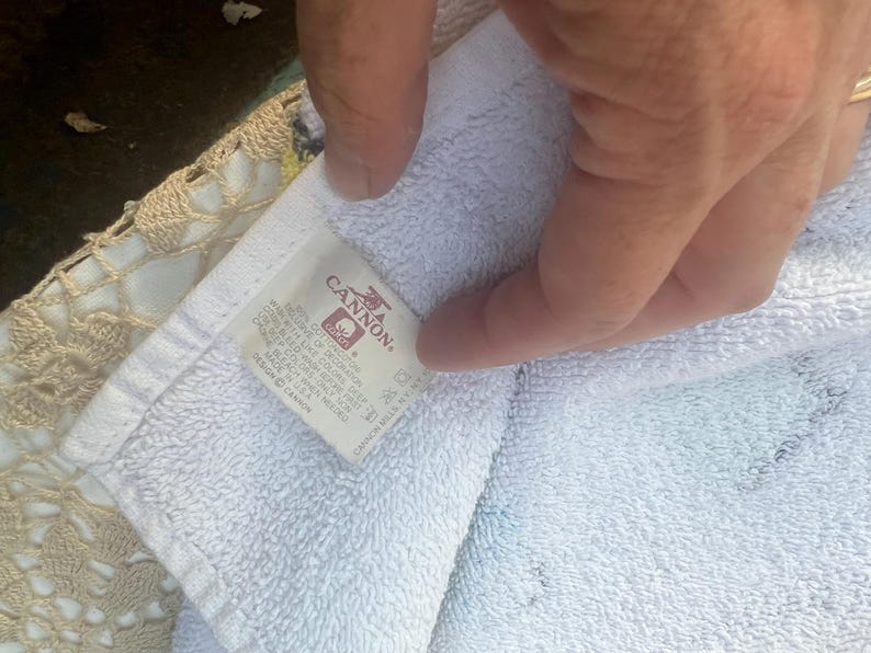 May include: White Cannon brand towel with a label visible. The label has the Cannon logo and text. The towel is folded and held by a hand. The towel is on top of a beige crocheted surface.