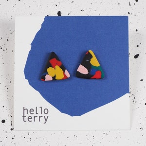 May include: A pair of triangular stud earrings with a black base and colorful abstract design. The earrings are displayed on a white card with a blue background and the text "hello terry". The background is white with black paint splatters.