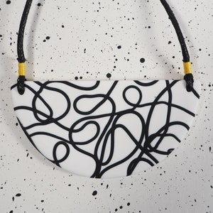 May include: A white, semi-circular pendant necklace with a black abstract design. The pendant is suspended from a black cord with yellow accents. The background is white with black paint splatters.