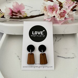 Puede incluir: Un par de pendientes de madera con tapas redondas negras. Los pendientes están en una tarjeta blanca con el texto "LOVE designs by george" y una dirección de sitio web.
