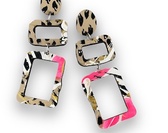 May include: A pair of dangle earrings with a geometric design. The earrings are made of wood and feature a leopard print, black and white stripes, and pink and white paint splatters.
