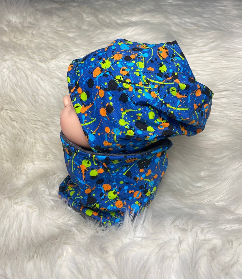 May include: Blue beanie and neck warmer set with a paint splatter pattern in black, orange, green, and yellow.