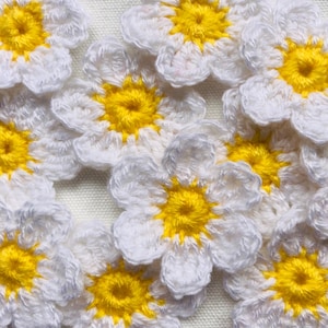 May include: A collection of crocheted daisy flowers with white petals and yellow centres. The flowers are arranged in a random pattern on a white background.