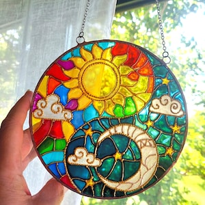 May include: A round, stained glass suncatcher featuring a sun, moon, clouds, and stars design. The sun is yellow, with a rainbow of colors surrounding it. The moon is white, and the background is teal. The suncatcher is hanging from a chain.