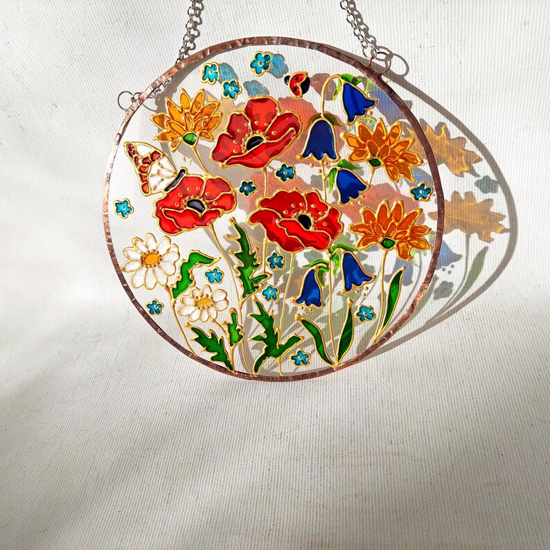 May include: A round stained glass panel featuring a colorful floral design. The artwork includes red poppies, bluebells, daisies, and a butterfly, all outlined in gold. The panel is suspended by a silver chain.