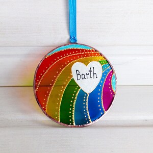 May include: A round, colorful glass ornament with a rainbow design and a white heart with the name "Barth" written on it.