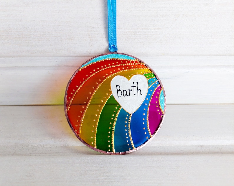 May include: A round, colorful glass ornament with a rainbow design and a white heart with the name "Barth" written on it.
