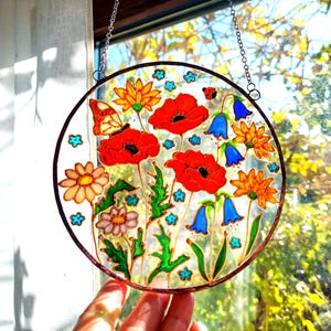 May include: A round stained glass window featuring a vibrant floral design. Red poppies, orange and yellow flowers, and blue bellflowers are depicted, along with a butterfly and ladybug. The artwork is framed with a brown border and hangs from a chain.