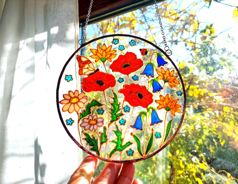 May include: A round stained glass window featuring a vibrant floral design. Red poppies, orange and yellow flowers, and blue bellflowers are depicted, along with a butterfly and ladybug. The artwork is framed with a brown border and hangs from a chain.