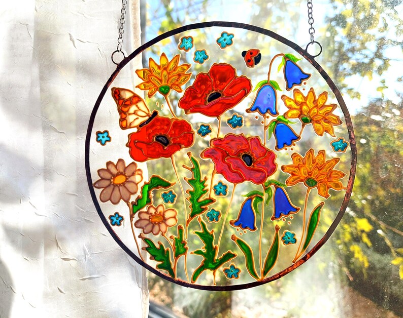 May include: A round stained glass panel featuring a vibrant floral design. The artwork includes red poppies, bluebell flowers, and orange daisies, with a butterfly and ladybug. The panel is framed with a black border and hangs from a chain.