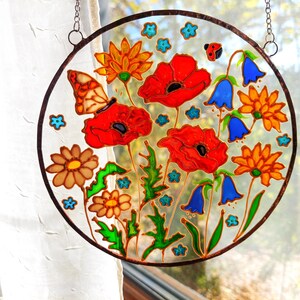 May include: A round, stained-glass window art piece featuring a vibrant floral design. The artwork showcases red poppies, orange and brown daisies, blue bellflowers, and a butterfly. The piece is framed with a dark brown border and hangs from a chain.
