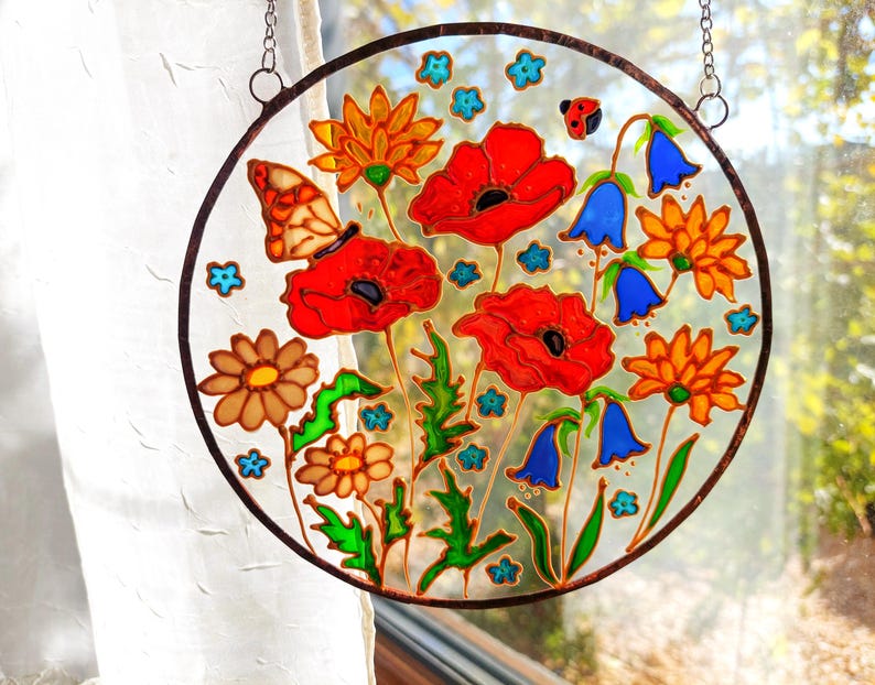May include: A round, stained-glass window art piece featuring a vibrant floral design. The artwork showcases red poppies, orange and brown daisies, blue bellflowers, and a butterfly. The piece is framed with a dark brown border and hangs from a chain.