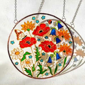 May include: Round stained glass panel featuring a vibrant floral design. The artwork showcases red poppies, orange and yellow flowers, bluebells, and a butterfly. The panel is framed with a copper-colored border and includes a hanging chain.