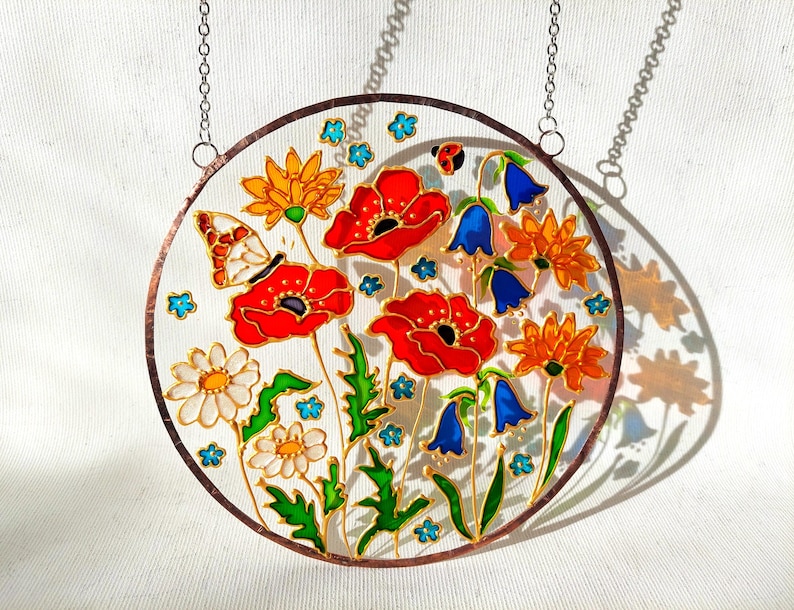 May include: Round stained glass panel featuring a vibrant floral design. The artwork showcases red poppies, orange and yellow flowers, bluebells, and a butterfly. The panel is framed with a copper-colored border and includes a hanging chain.
