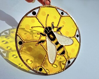 Può includere: Suncatcher a forma di ape in vetro colorato con un design a nido d'ape. L'ape ha un corpo nero e giallo con ali bianche. Il nido d'ape è giallo con punti neri. Il suncatcher è rotondo con una cornice in metallo color rame.
