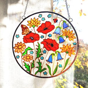 May include: A round stained glass window featuring a colorful floral design. The artwork includes red poppies, yellow daisies, bluebell flowers, and a butterfly. The glass is framed with a dark border and suspended by a chain.