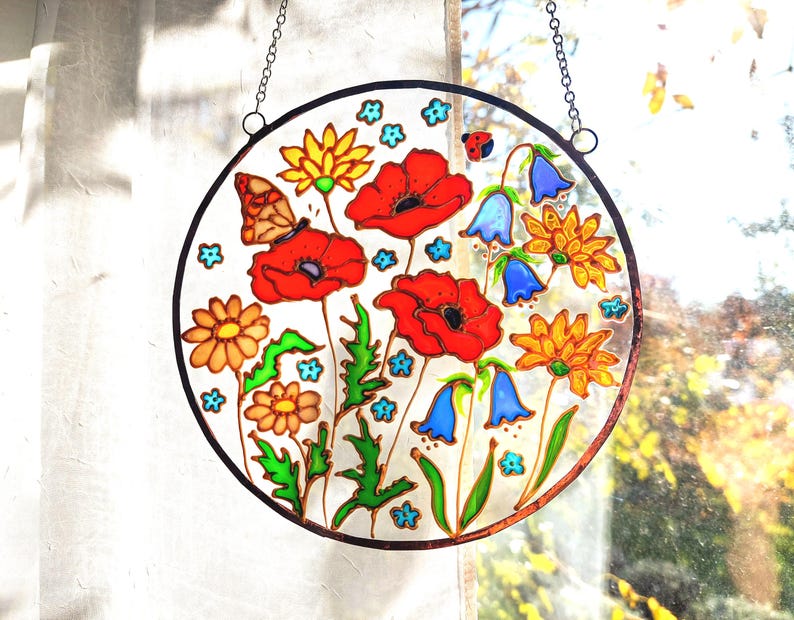 May include: A round stained glass window featuring a colorful floral design. The artwork includes red poppies, yellow daisies, bluebell flowers, and a butterfly. The glass is framed with a dark border and suspended by a chain.